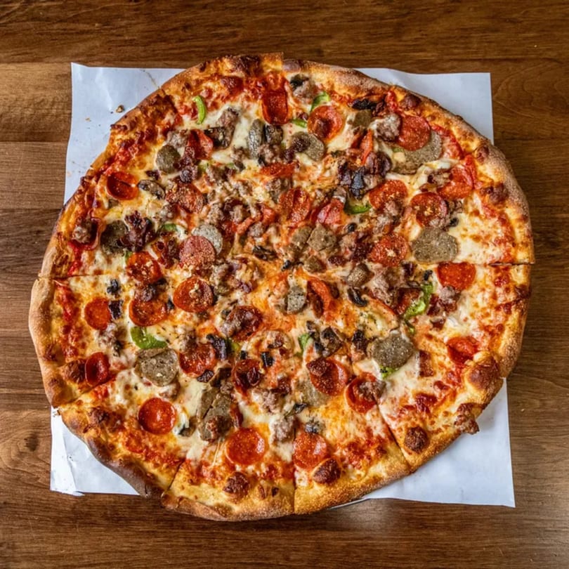 A whole Pesto's pizza, topped with pepperoni, sausage, mushrooms, and green peppers, on a wooden table.