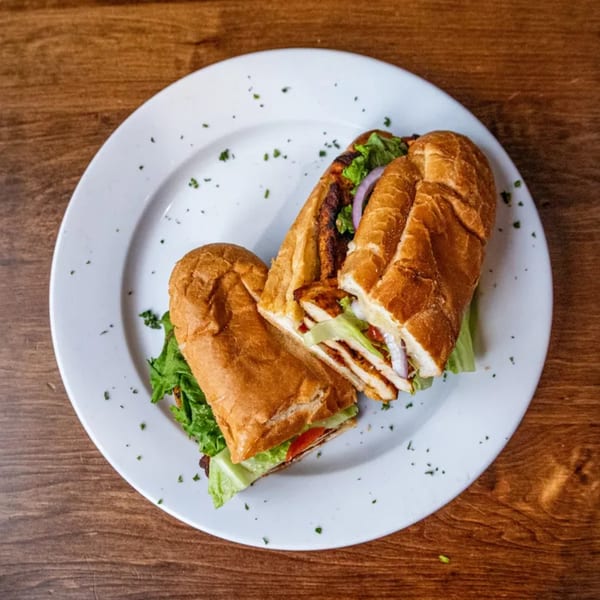 A Pesto's Italian chicken sub sandwich, cut in half, on a white plate with lettuce, tomato, and onion.