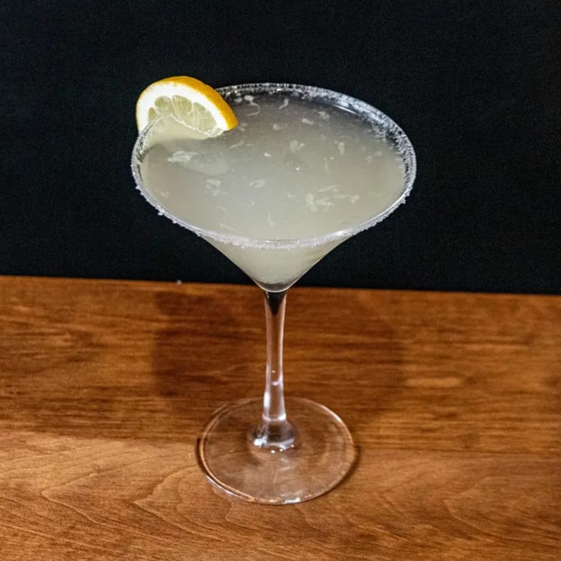 A Pesto's lemon drop martini, garnished with a lemon slice and sugar rim, on a wooden table.